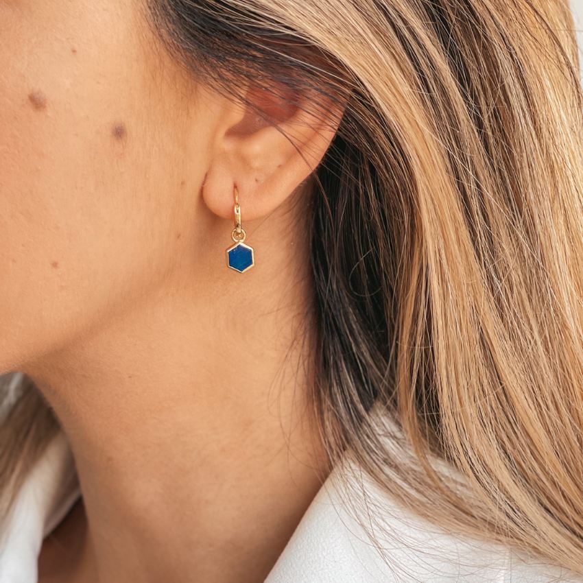 A close-up of a woman's ear wearing a golden earring with a blue hexagonal stone, identified as Lapis Lazuli.