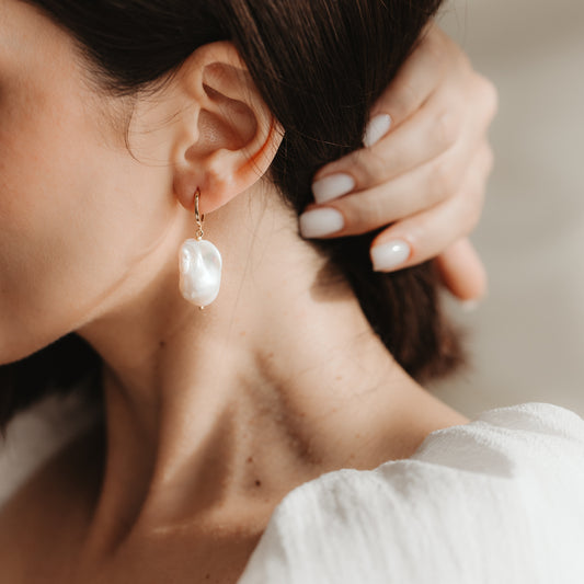 Close-up of an ear wearing a gold-plated 925 silver hoop earring with a large white baroque pearl; hand with neutral manicure near the ear in soft natural light.