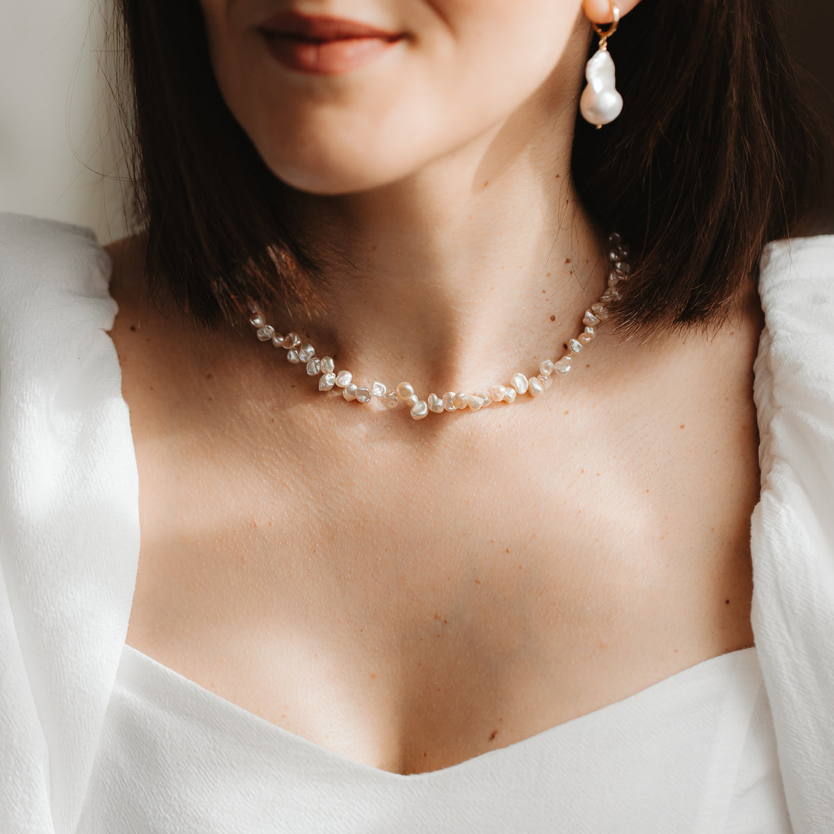 Close-up of a model wearing a delicate pink keshi pearl necklace and a gold-plated baroque pearl drop earring, styled with a white dress in soft natural light.