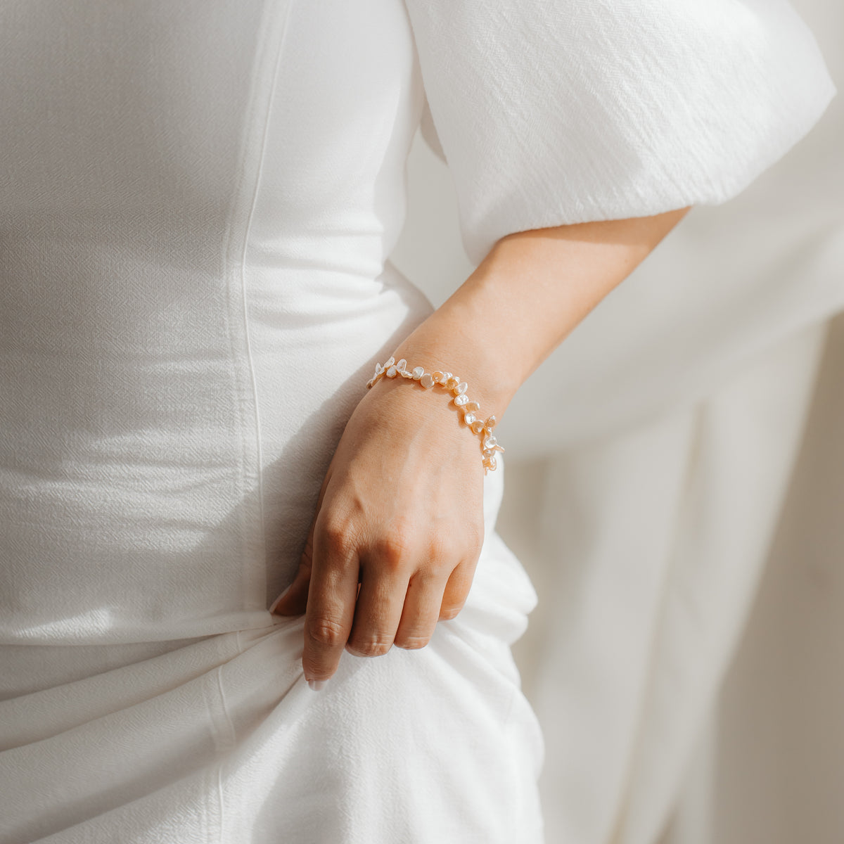 Close-up of a gold‑plated sterling silver bracelet with pink keshi pearls on a person's wrist, styled against a white dress in soft light.