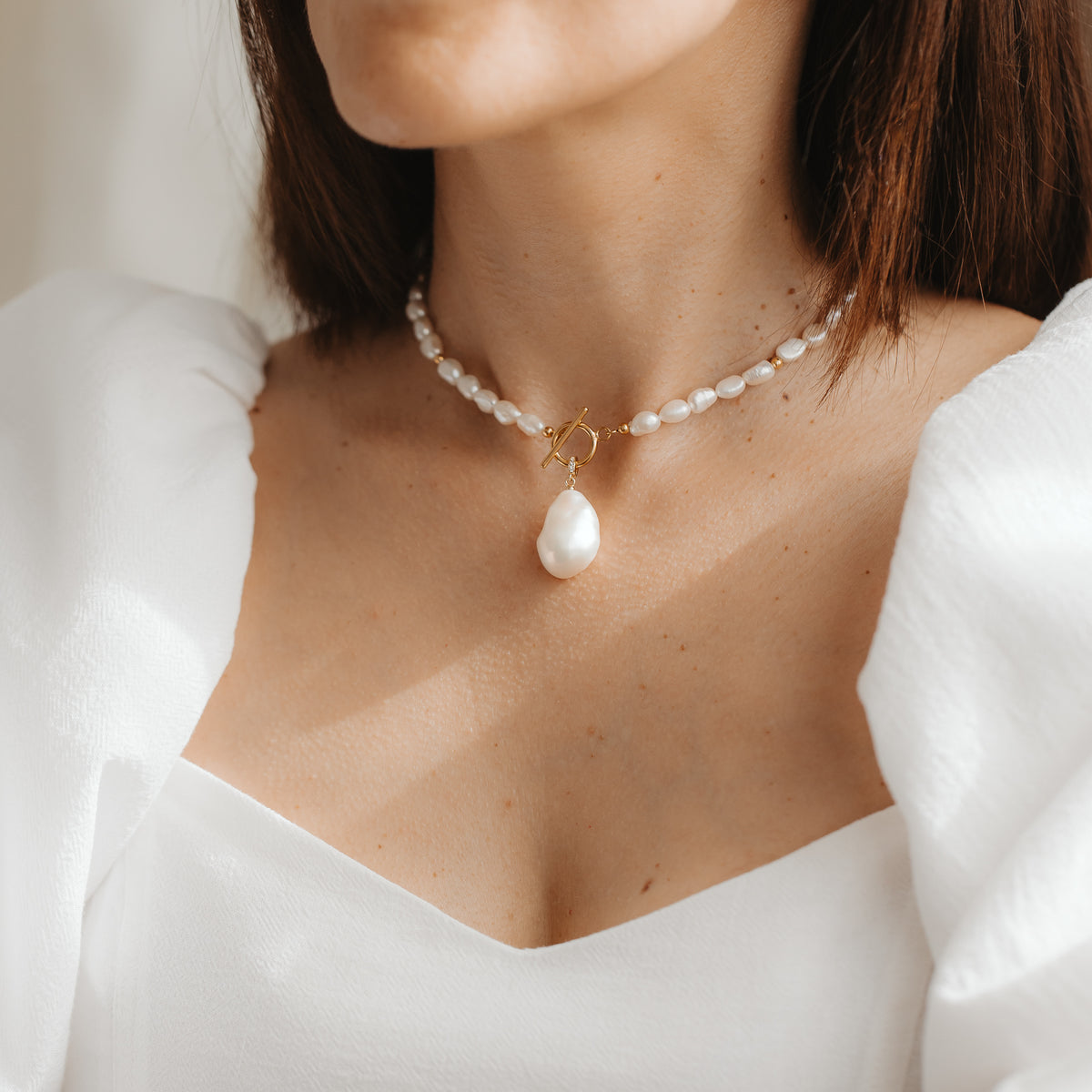 Close-up of a woman wearing a short natural pearl necklace with a baroque pearl pendant and a gold toggle clasp, styled with a white dress.