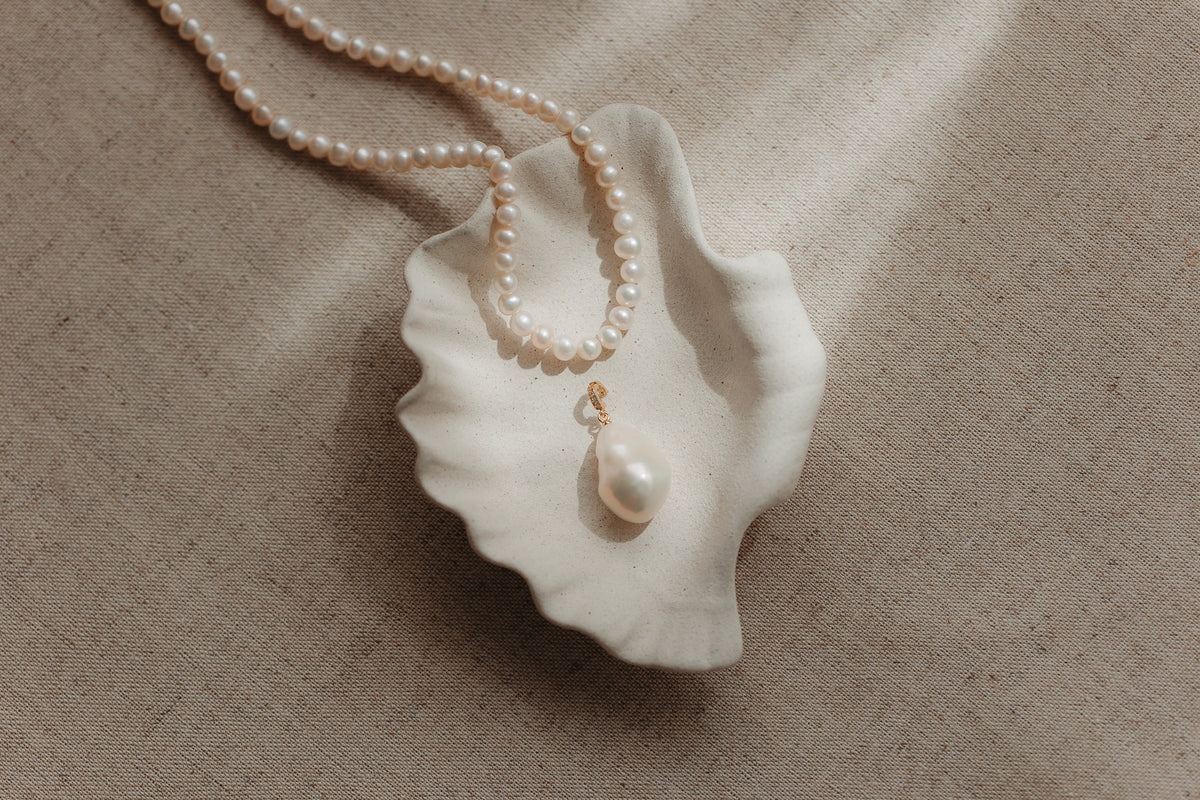 Baroque pearl pendant with a gold‑plated silver bail resting in a scalloped ceramic shell dish, with a pearl strand on a beige linen background.