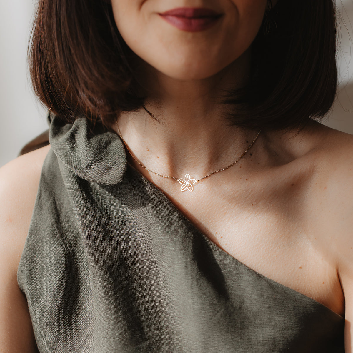 Close-up of a person wearing a delicate 14K yellow gold wildflower outline necklace at the collarbone, styled with an olive one-shoulder top in natural light.