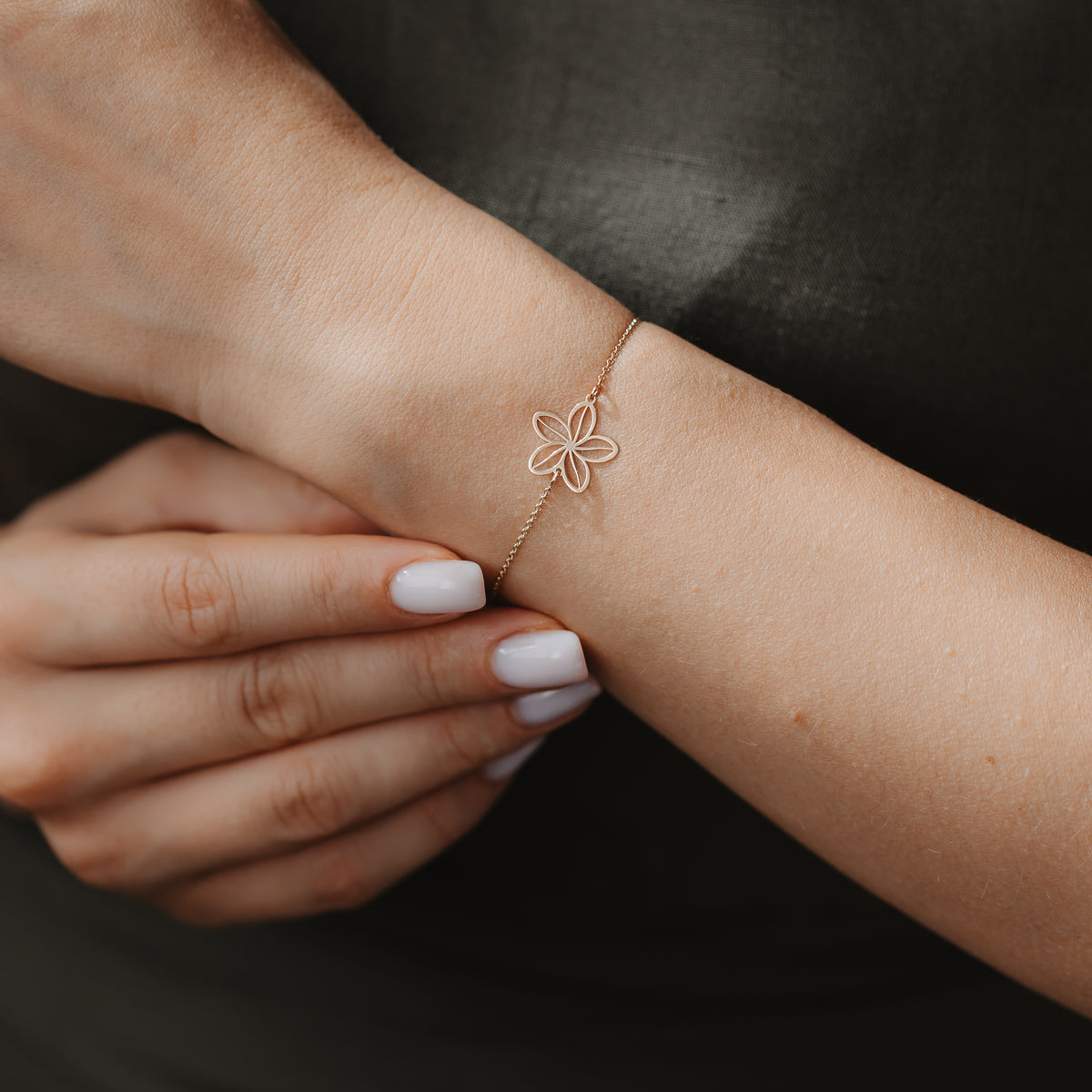 Close-up of a delicate 14K gold bracelet with an openwork wildflower charm on a wrist, against a dark fabric background.