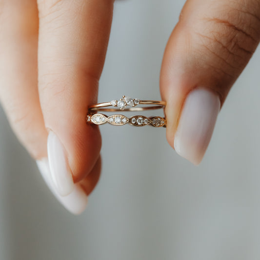 Close-up of two 14K yellow gold diamond stacking rings held between fingers—one with a petite cluster and one with marquise-shaped stations.
