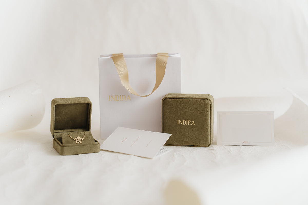 Indira gift packaging set: white shopping bag with gold ribbon handles, two olive velvet jewelry boxes (one open showing a gold flower pendant), and branded cards on a soft white studio background.