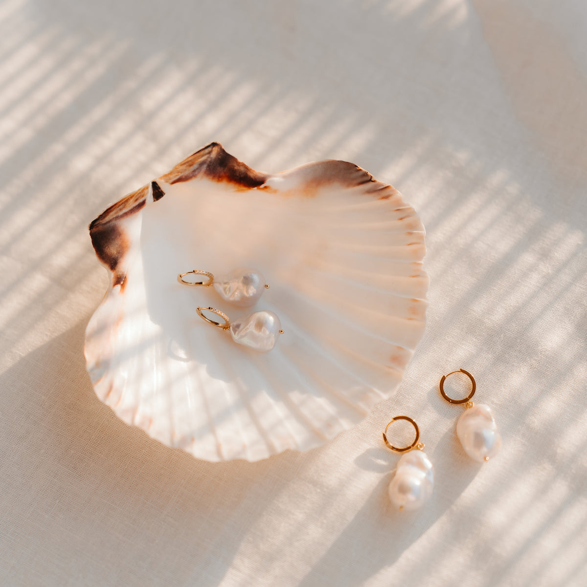 Baroque pearl drop earrings with gold‑plated sterling silver hoops and zirconia accents styled on a scallop shell over linen in warm daylight.