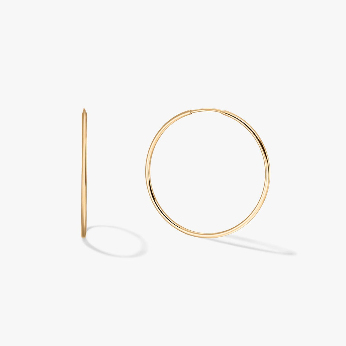 Pair of large thin 14K yellow gold hoop earrings on a white background, one shown front-on and one in profile with soft shadows.