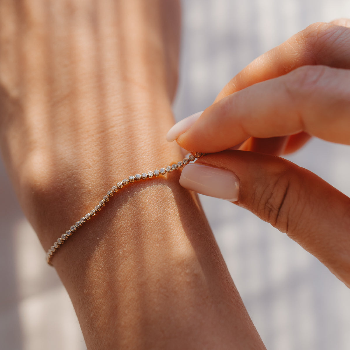 Close-up of a hand fastening a 14K yellow gold diamond tennis bracelet on a wrist in soft sunlight.