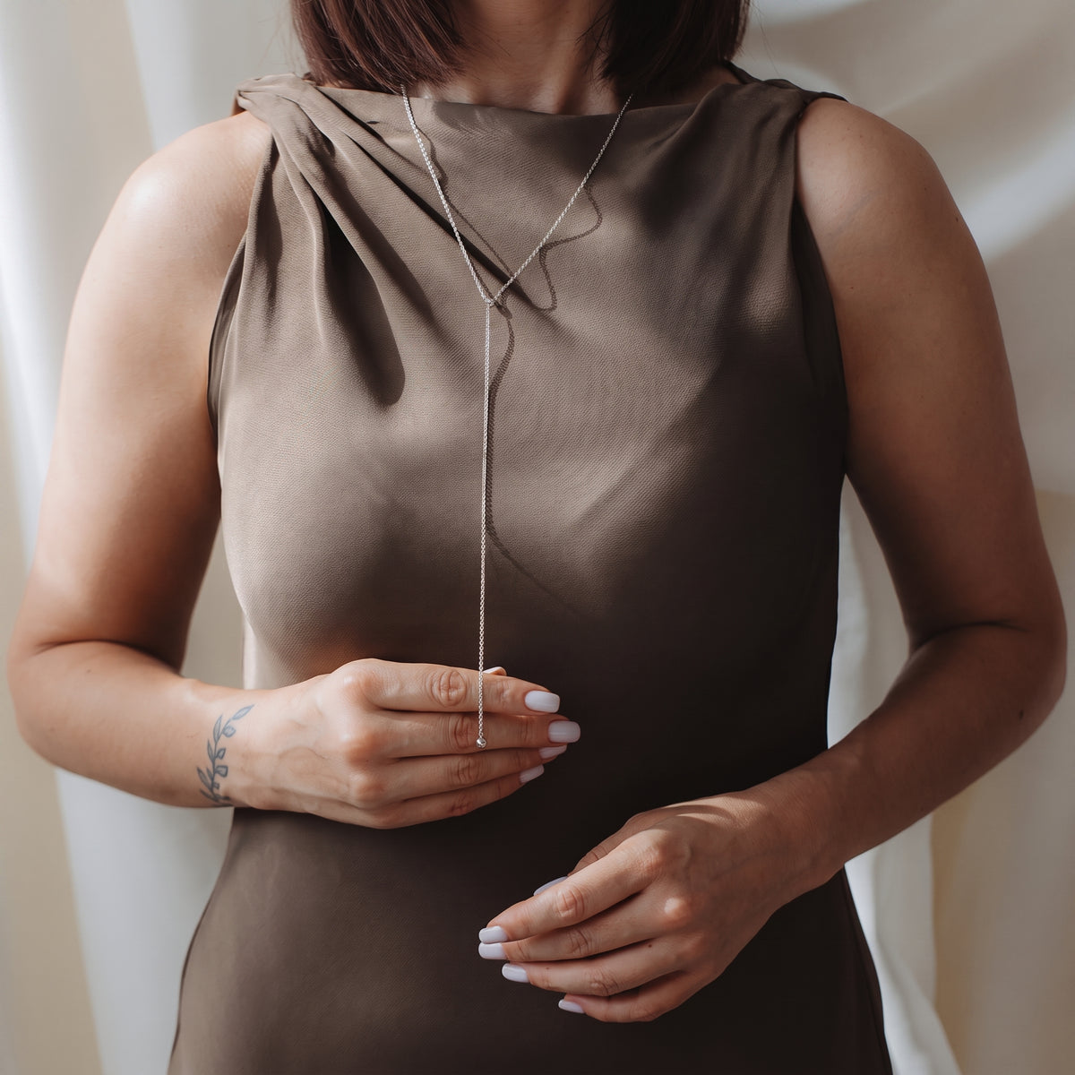 Close-up of a model wearing a 925 silver bola lariat necklace over a taupe sleeveless dress, lightly holding the drop chain.