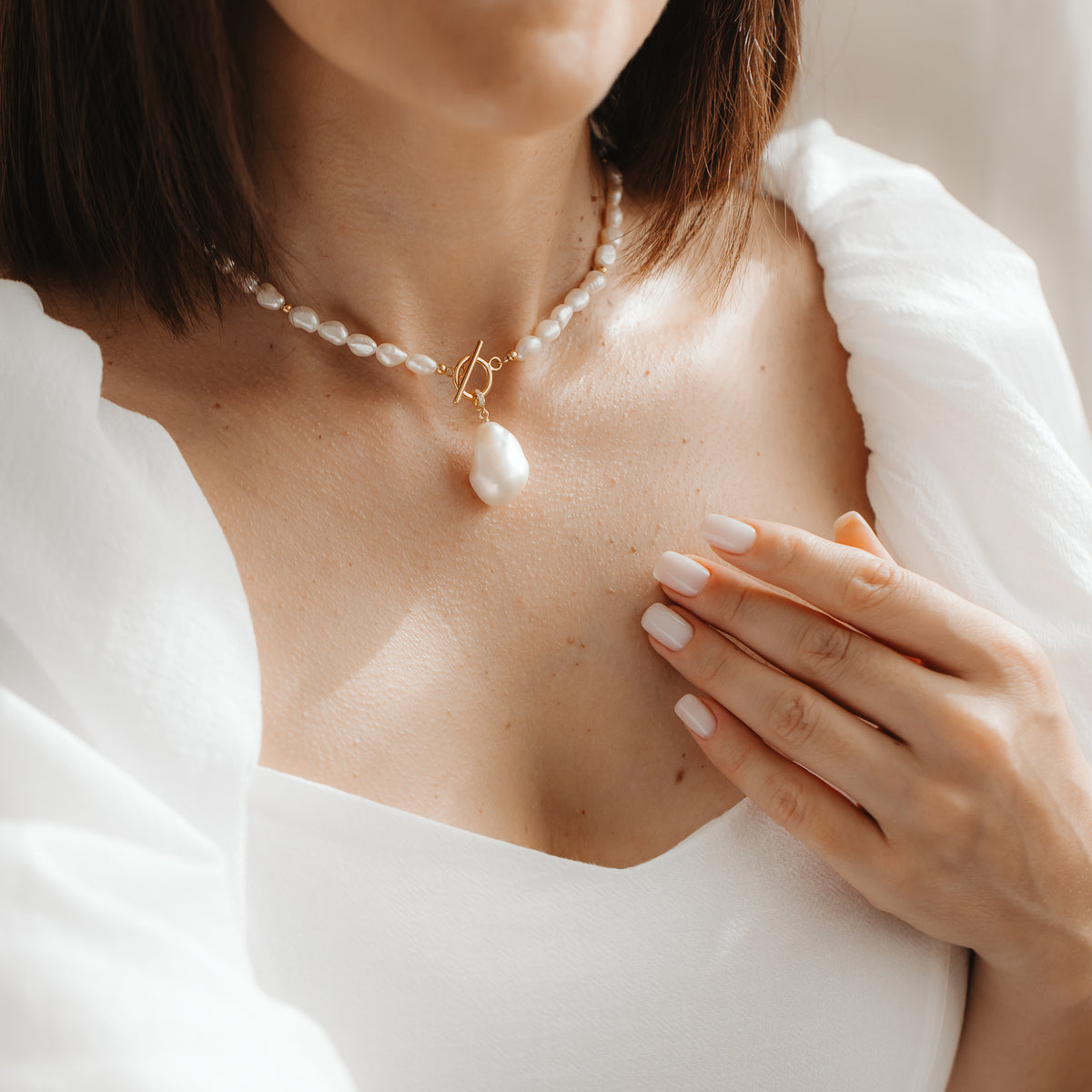 Close-up of a woman wearing a strand of natural pearls with a gold‑plated baroque pearl pendant, touching her collarbone in soft daylight.