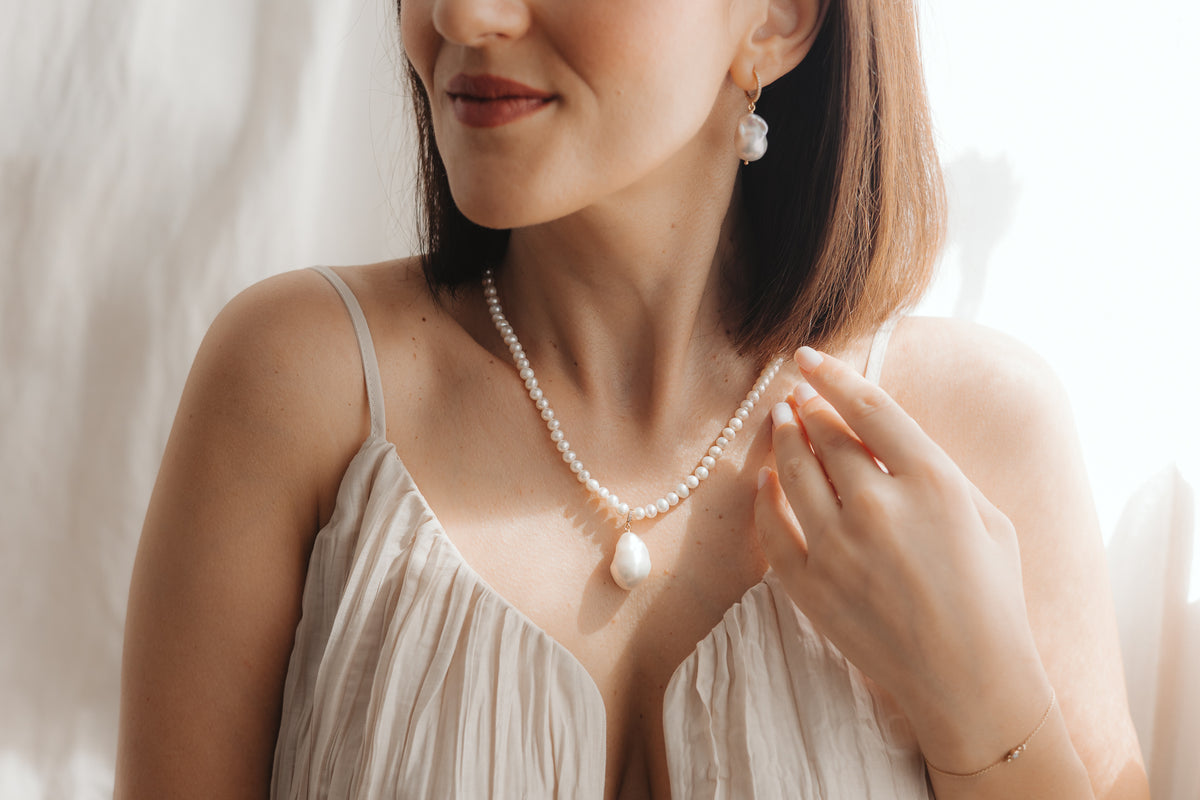 Close-up of a woman wearing a baroque pearl pendant on a pearl necklace with a matching gold-plated silver drop pearl earring, lit by soft natural light.