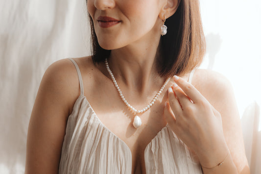 Close-up of a woman wearing a baroque pearl pendant on a pearl necklace with a matching gold-plated silver drop pearl earring, lit by soft natural light.