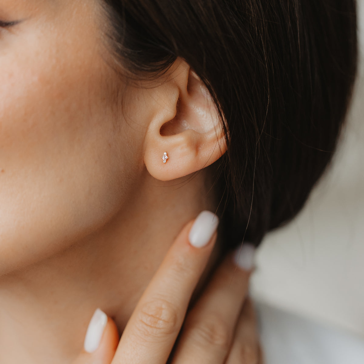 Close-up of an ear wearing a tiny 14K yellow gold Flora stud earring with pink morganite and white sapphire, while a hand with a neutral manicure touches the neck.