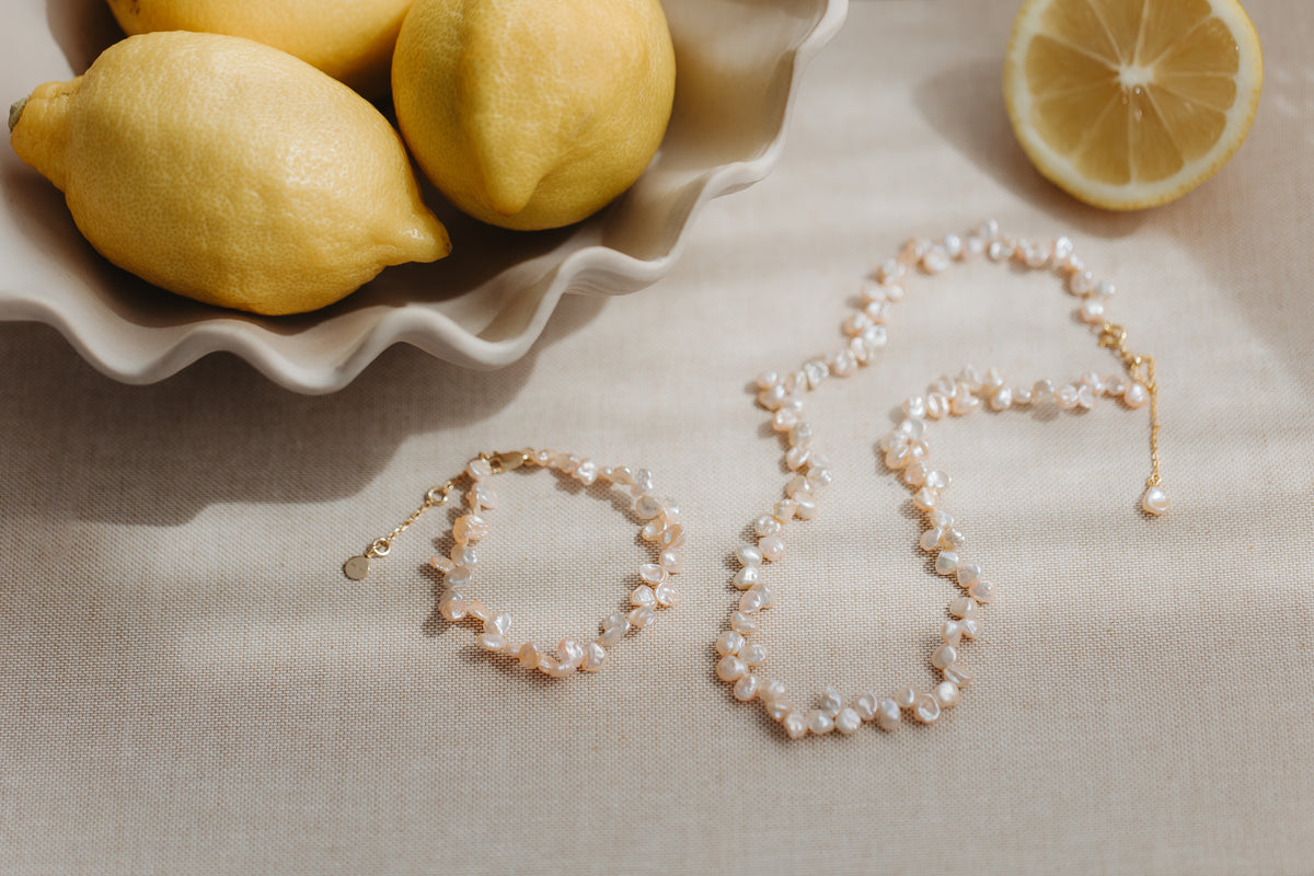 Pink keshi pearl necklace and matching gold‑plated silver bracelet styled on beige linen with fresh lemons.