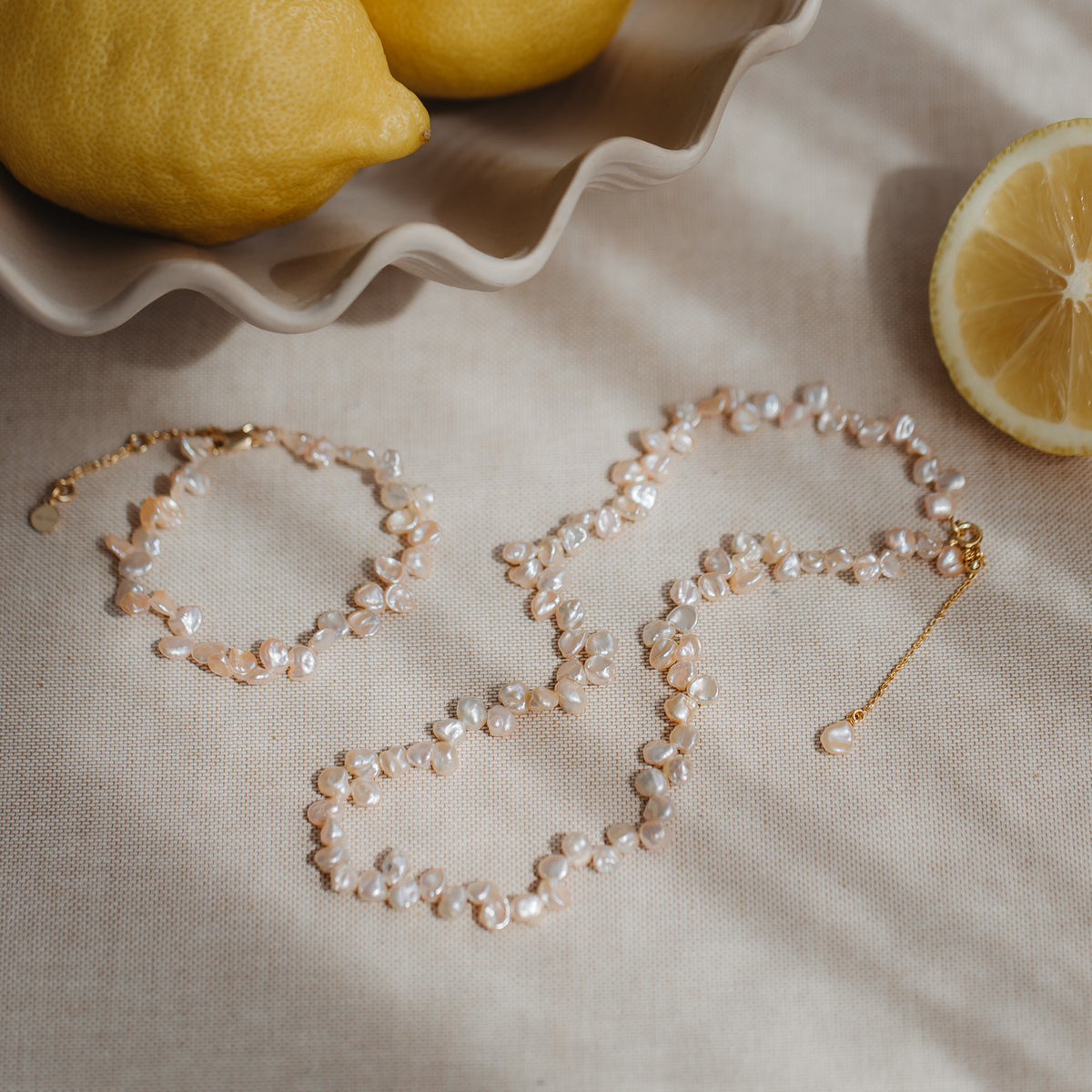Gold‑plated silver necklace with pink keshi pearls styled on neutral linen with lemons, with a matching bracelet nearby.