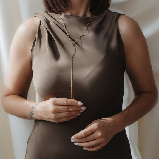 Close-up of a person wearing a gold‑plated 925 silver lariat (Bola) necklace over a taupe satin dress, with hands gently adjusting the drop pendant.