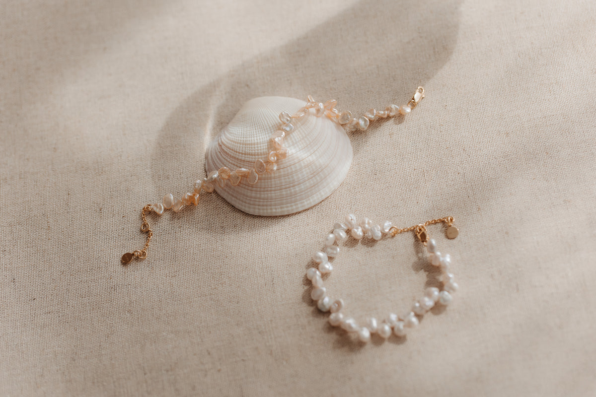 Two gold‑plated sterling silver bracelets with pink keshi pearls styled on a seashell over beige linen fabric.