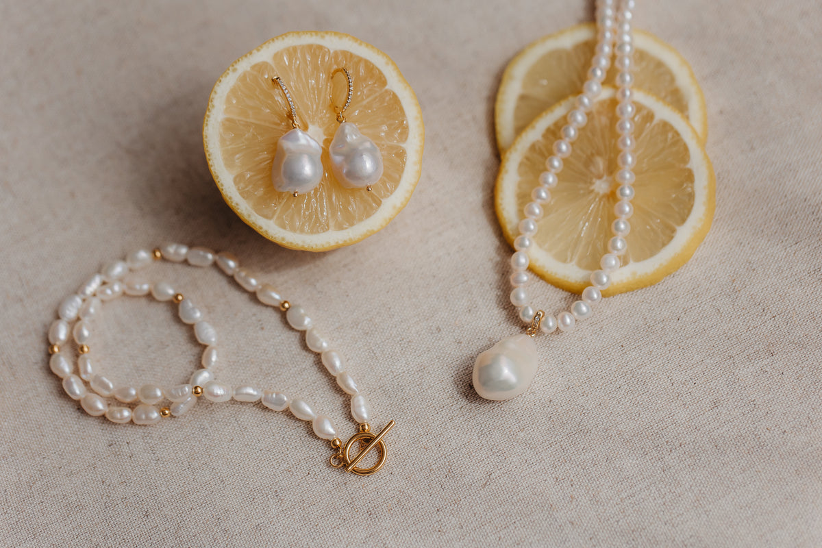 Baroque pearl earrings on gold-plated sterling hoops pinned into a lemon half, with matching pearl necklace and bracelet styled on beige linen.