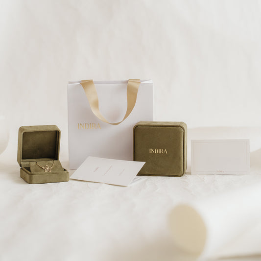 Indira branded gift packaging set with a white bag and gold ribbon handles, olive velvet jewelry boxes (one open), and cards arranged on a soft neutral background.