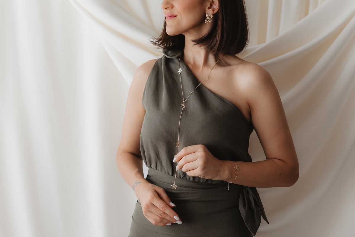 Close-up of a model wearing the Wildflower Maxi 14K yellow gold lariat necklace with delicate flower charms, styled over an olive one-shoulder dress against a soft draped cream backdrop.