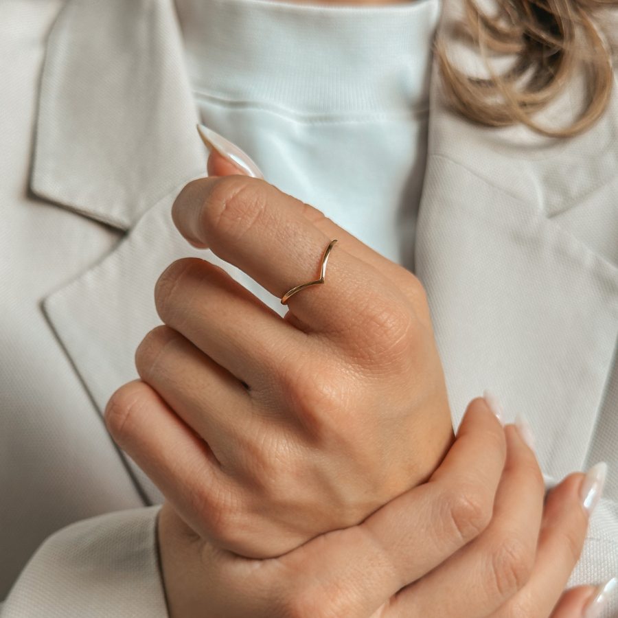 A person wearing a simple V-shaped golden ring on their finger, dressed in a light suit.