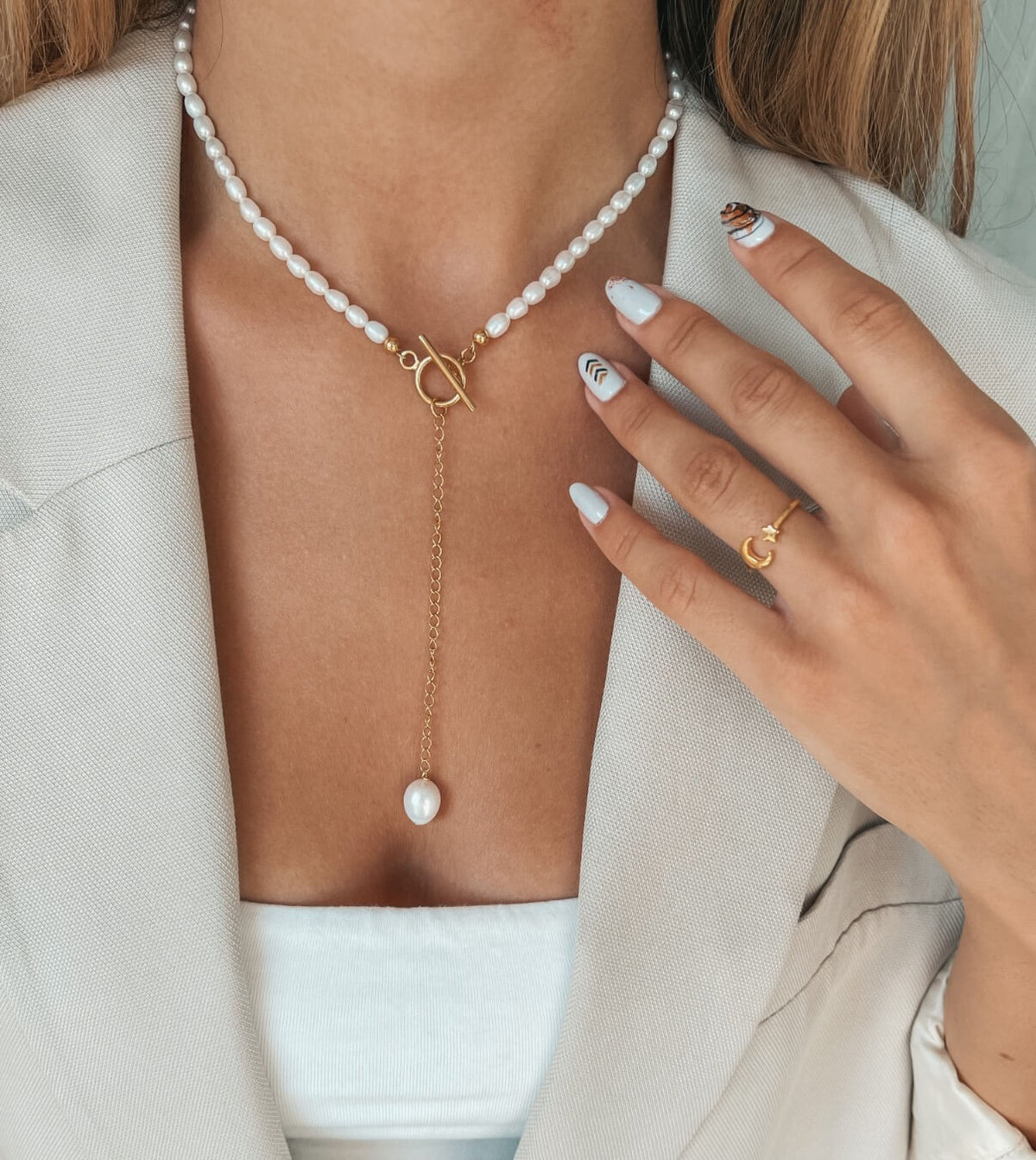 Close-up of a woman's neck adorned with a pearl necklace featuring a gold chain and a hanging pearl, complemented by a minimalist gold ring on her finger.